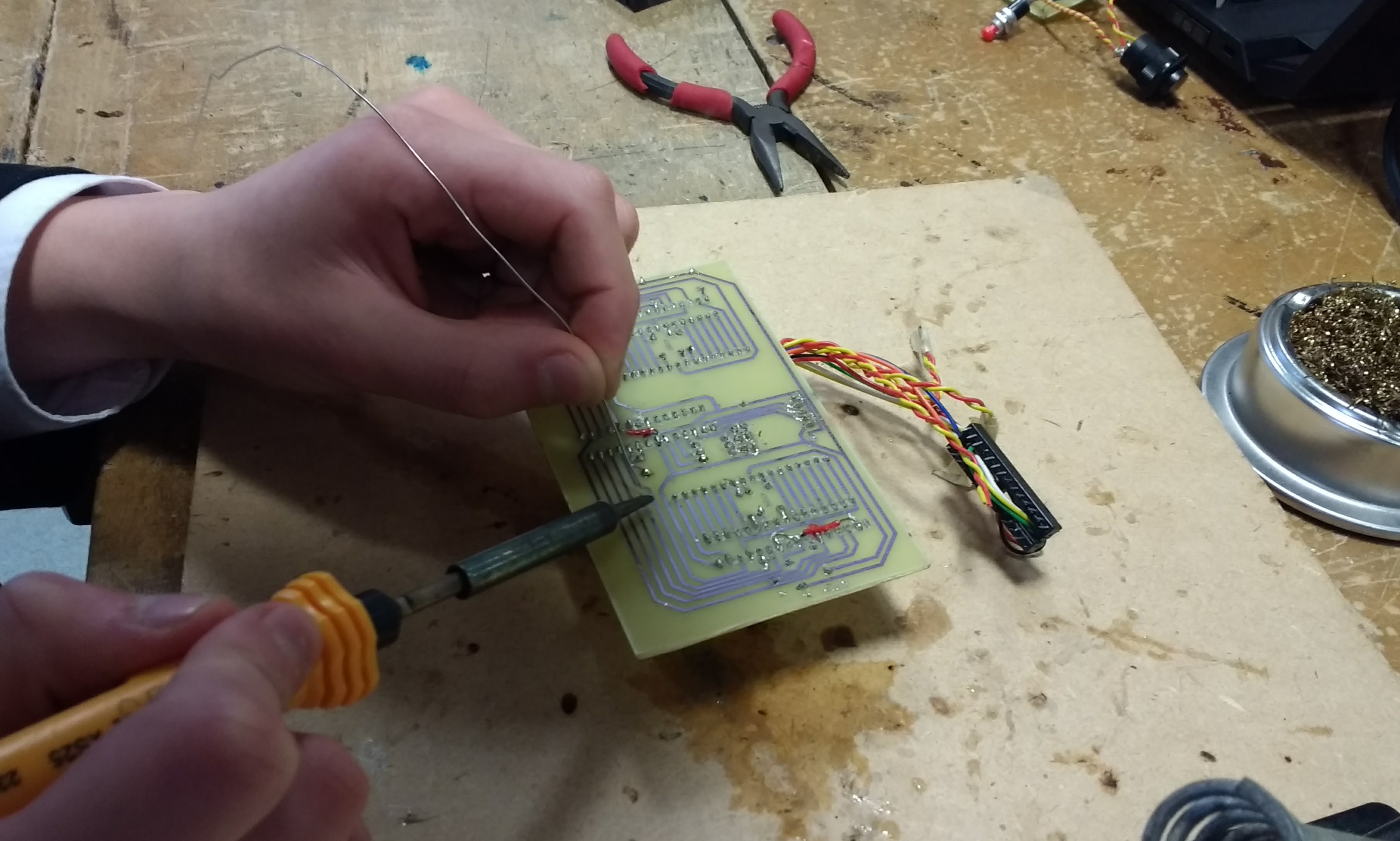 Teenage hands soldering a hand-etched circuit board at a workbench: soldering iron in one hand, a component lead in the other, side cutters and a solder reel on the bench