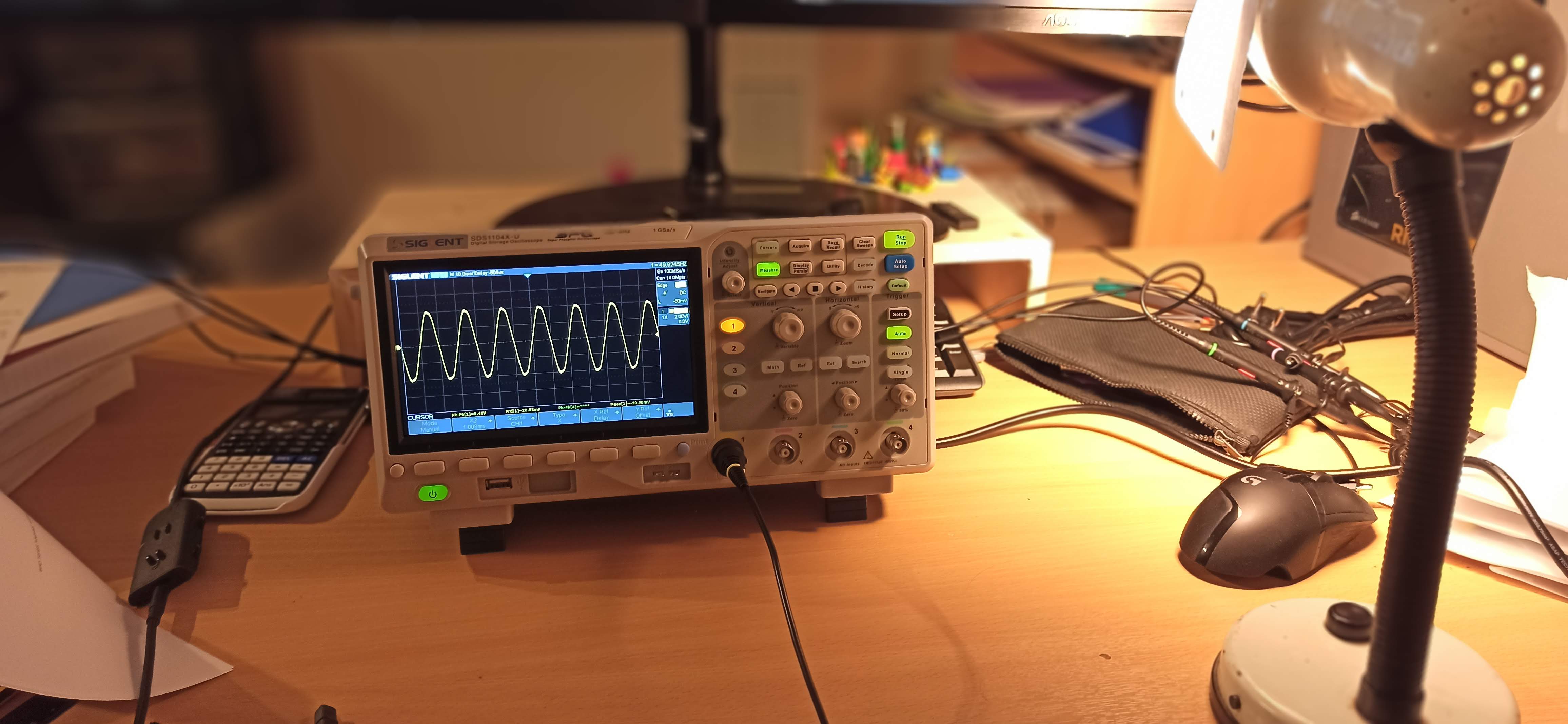 Siglent oscilloscope displaying a sine wave on a cluttered desk beside a calculator, lamp and monitor